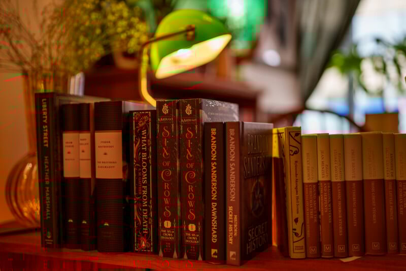 A row of assorted hardcover and paperback books on a wooden shelf, illuminated by a green desk lamp, with blurred plants and warm lighting in the background, creating a cozy reading atmosphere.