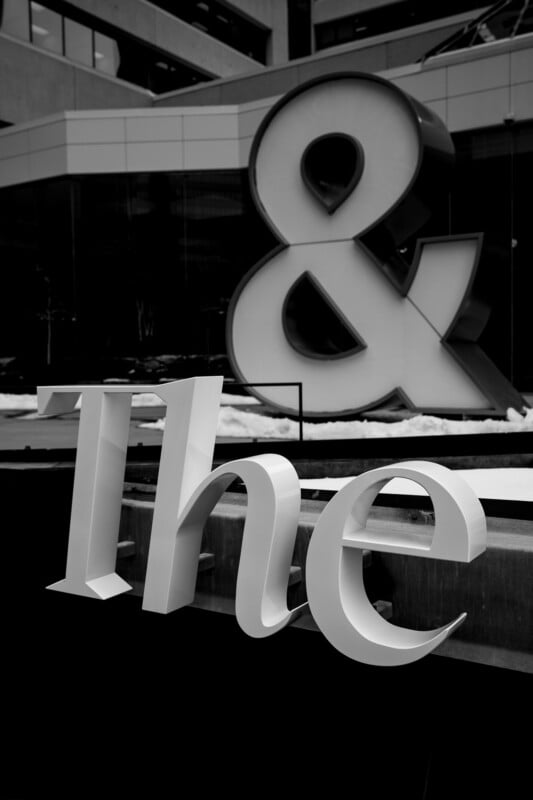 Large white letters spelling "The" are in the foreground, with a giant ampersand (&) sculpture in the background, both set against a modern building in a black and white photo.