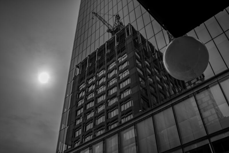 Black and white photo of a modern glass building reflecting another building with a crane on top, with the sun shining through a cloudy sky and a round object in the upper right foreground.