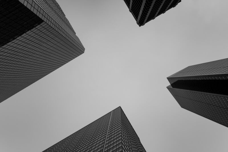 Black and white photo looking up at four tall skyscrapers with glass windows, set against a cloudy sky, creating a dramatic and geometric perspective.