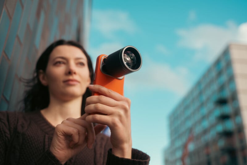 A woman holds a smartphone with an attached external lens, aiming it upwards outdoors. She stands near a modern building with a blue sky and clouds in the background.