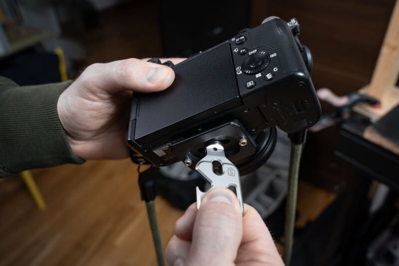 A person uses a metal tool to tighten or loosen a screw on the bottom of a black digital camera, holding the camera in one hand and the tool in the other.