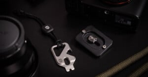 A close-up of a metal multi-tool with a black cord, a camera quick-release plate, a camera body, and a camera lens, all arranged on a dark surface. The multi-tool features a logo and multiple functions.
