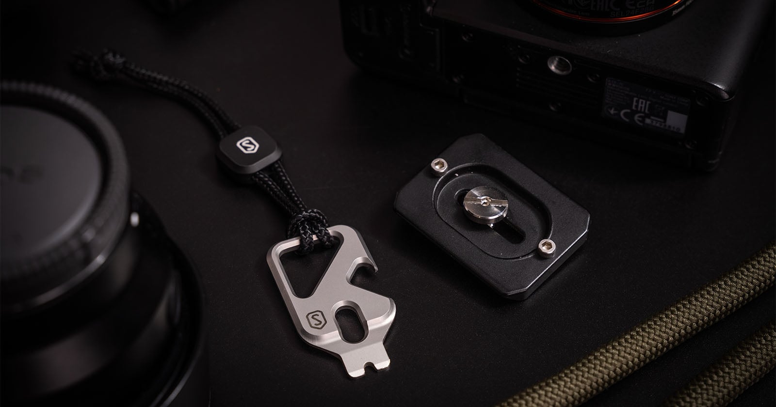 A close-up of a metal multi-tool with a black cord, a camera quick-release plate, a camera body, and a camera lens, all arranged on a dark surface. The multi-tool features a logo and multiple functions.