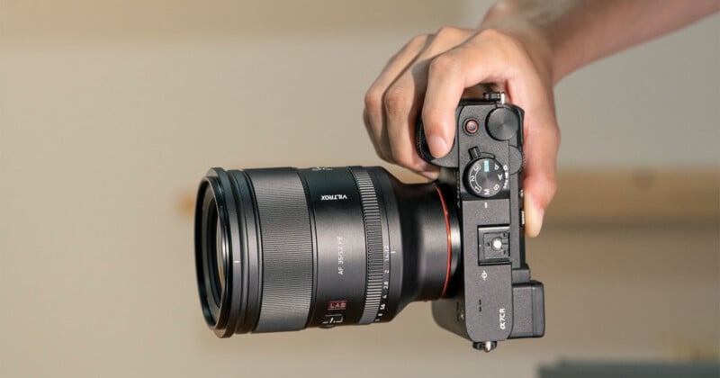 A close-up of a person’s hand holding a black digital camera with a large lens, pointed forward, against a plain background. The camera’s controls and dials are clearly visible.