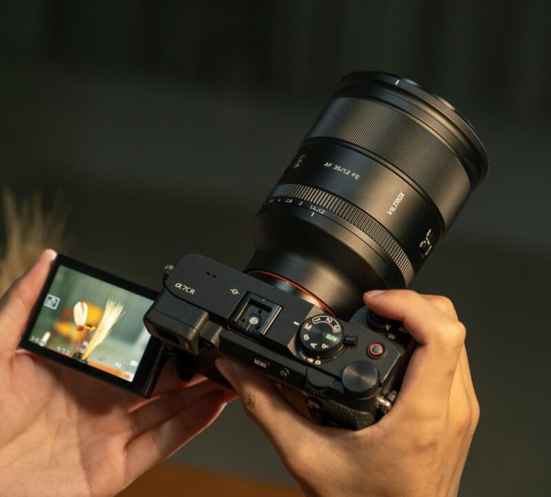 A person holds a digital camera with a large lens, adjusting settings. The camera's flip-out screen displays a close-up image of a bird perched on a branch.