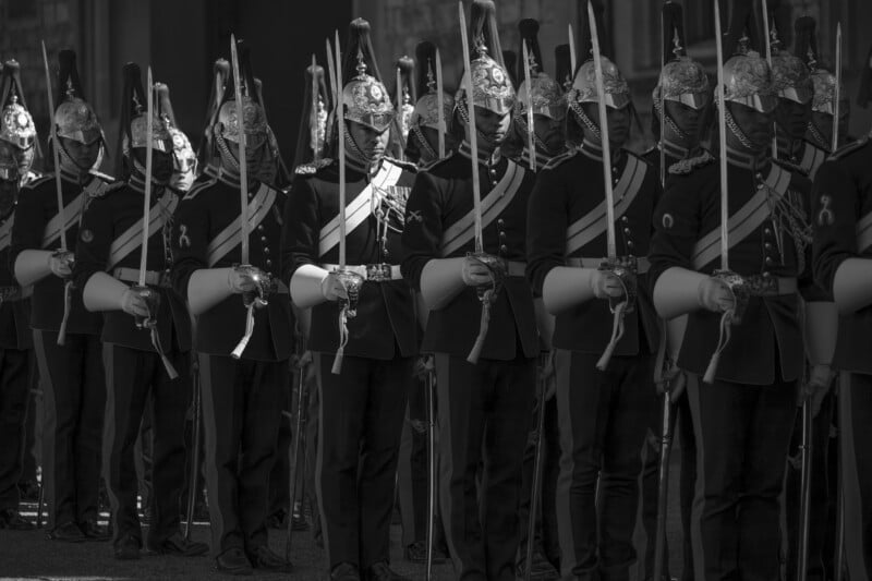 A row of ceremonial guards in ornate uniforms and shiny helmets stand in formation, holding swords vertically in front of them. The image is in black and white, with dramatic lighting highlighting their uniforms.