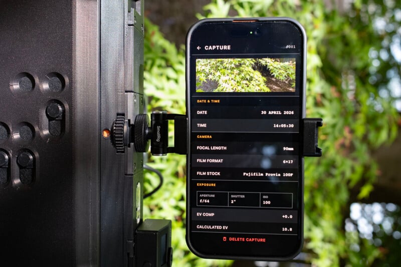A smartphone mounted on a camera rig displays image capture settings, including date, time, focal length, film format, film stock, exposure, and EV compensation, with green foliage visible in the background.