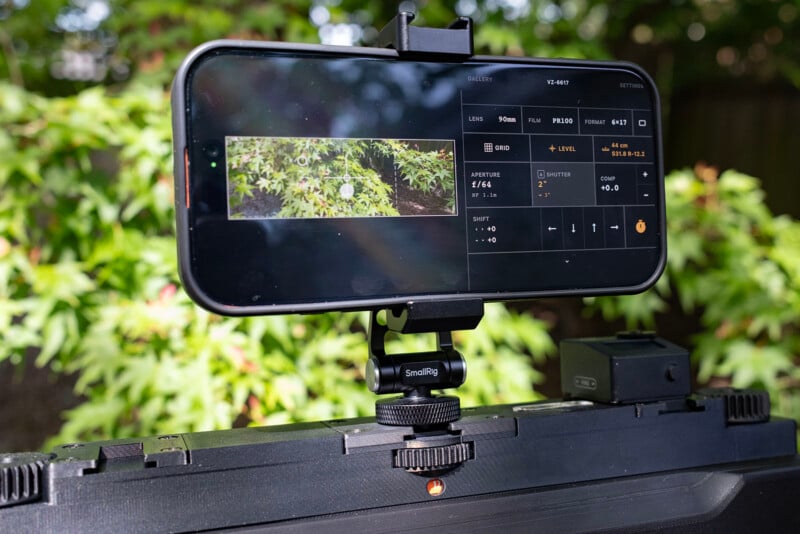 A smartphone mounted on a tripod records green leaves, displaying camera settings like ISO and shutter speed on its screen. The background features lush, sunlit foliage.