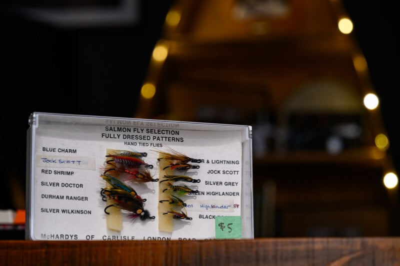 A clear box labeled “Salmon Fly Selection” displays colorful, hand-tied fishing flies. The box rests on a wooden surface with blurred fishing gear in the background and a green $5 price tag attached.