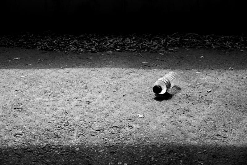 A plastic bottle lies on a rough, sunlit patch of ground, with dark shadows and scattered debris in the background.