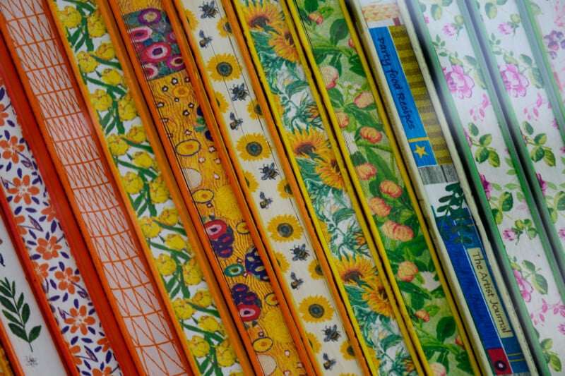 Rows of colorful book spines with vibrant floral and geometric patterns, including sunflowers, daisies, and leaves, are arranged vertically, showcasing a variety of decorative designs and textures.