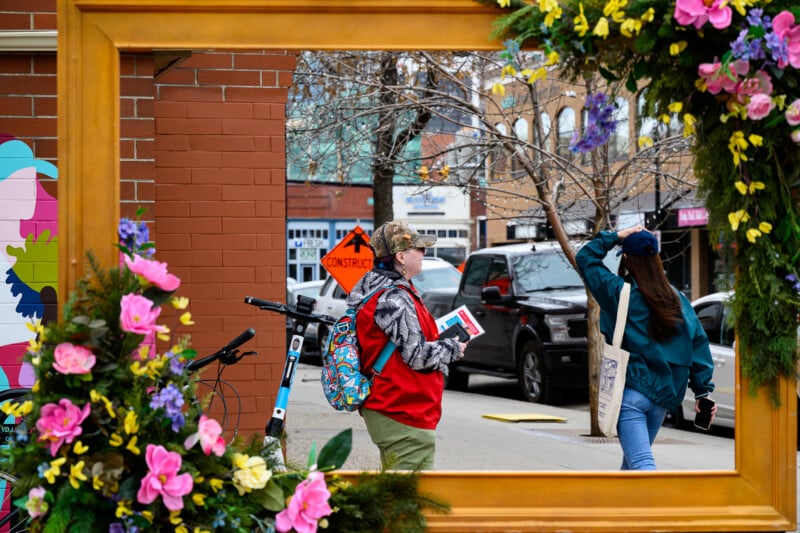 A person with a colorful backpack and red jacket stands next to a bicycle, framed by a large wooden frame decorated with flowers on a city sidewalk, while another person walks by on the street.