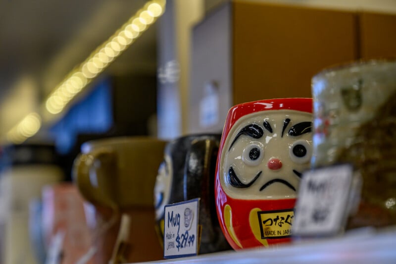 A close-up of a ceramic Daruma doll with a red body and black eyebrows on a store shelf, surrounded by other mugs and pottery, with a blurred background and price tags visible.