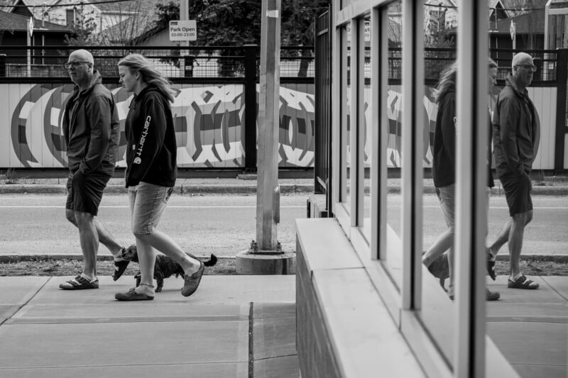 A man and woman walk on a sidewalk, reflected in the glass windows to their right. The scene is in black and white, with a patterned fence and a "Park Open" sign in the background.