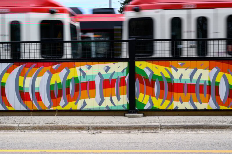 A colorful mural featuring geometric shapes and bold stripes decorates a fence along a sidewalk, with a red-and-white train passing by in the background.