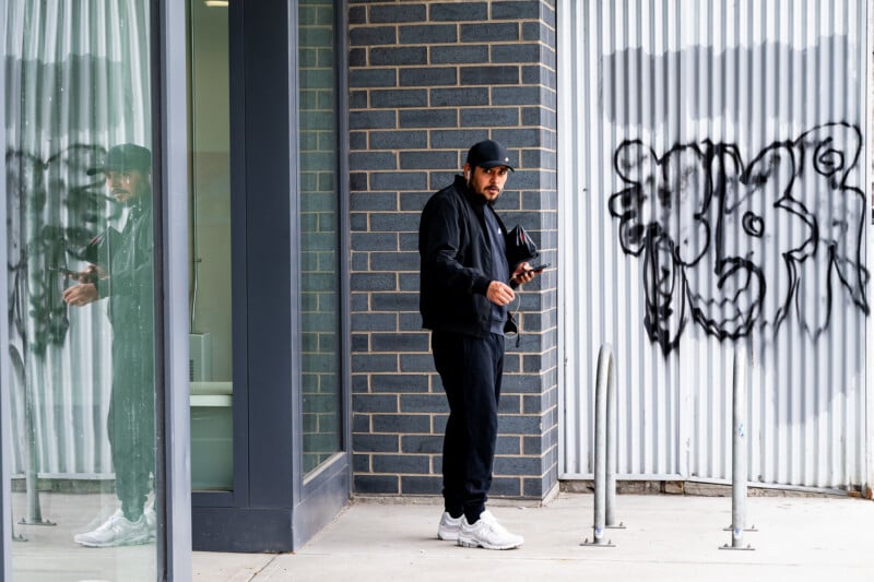 A man in black clothing and a cap stands outside a building with a phone in hand. His reflection appears on a glass wall. Behind him is graffiti on a corrugated metal wall and empty bike racks.