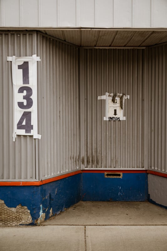 A corrugated metal wall with a large sign reading "134" on the left, a small box with tape and stickers in the center, and a weathered blue and red base along the sidewalk.