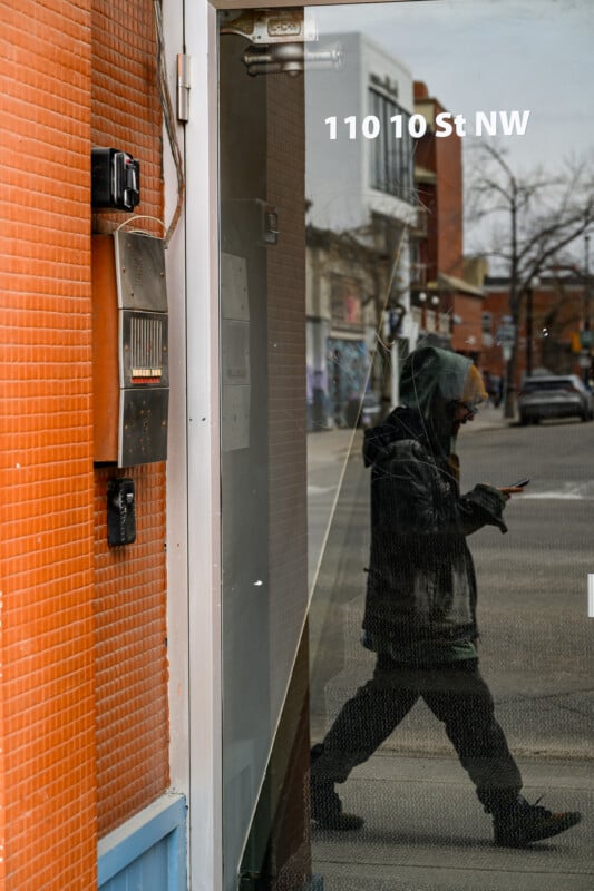 A person in a hooded jacket walks past a glass door at 110 10 St NW, their reflection visible in the glass as they look down at their phone; city buildings and a street are seen in the background.