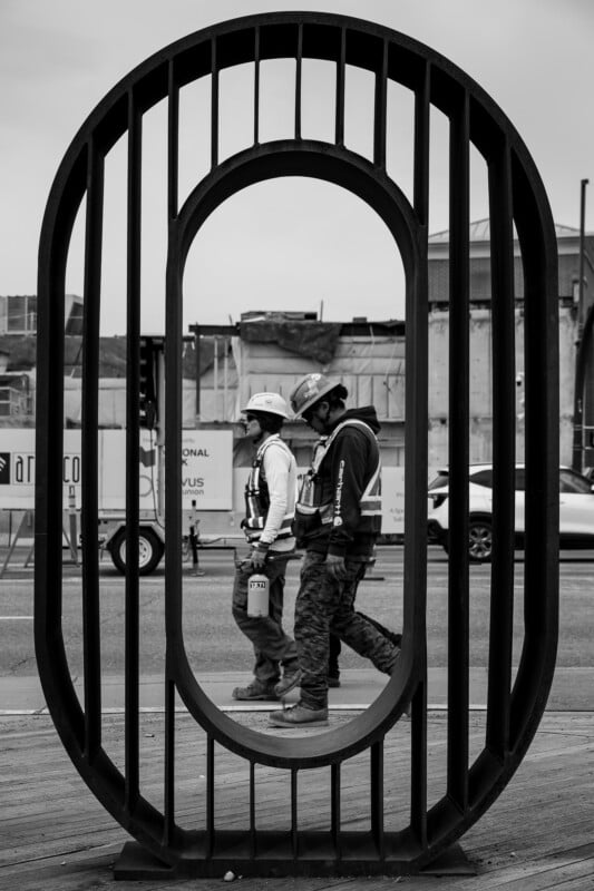 Two construction workers in safety gear walk side by side, framed by a large oval metal sculpture. The background shows urban buildings and vehicles. The photo is in black and white.
