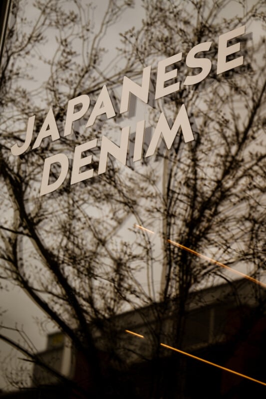 The image shows the words "JAPANESE DENIM" in bold white letters on a window, with the reflection of leafless tree branches and some building features visible in the glass.