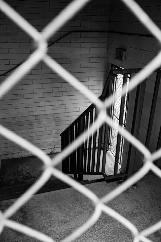 Black and white photo of a stairwell viewed through a chain-link fence; a door marked "EXIT" is open at the bottom of the stairs, with sunlight streaming inside.