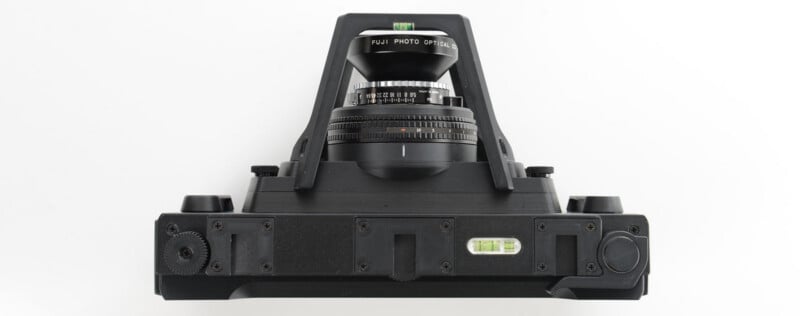 A top-down view of a black camera with a wide lens and two bubble spirit levels, set against a white background. The camera features dials, knobs, and a sturdy frame.