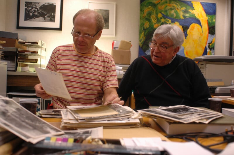 Two older men sit at a desk covered in photographs and papers, reviewing documents together in an office with artwork and folders in the background.