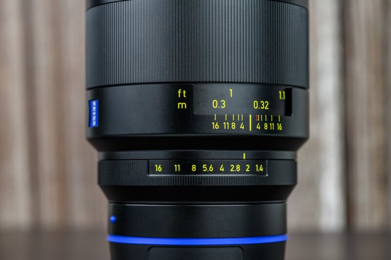 Close-up of a camera lens showing focus distance markings in feet and meters, and an aperture ring with f-stop values ranging from f/1.4 to f/16. The background is blurred wood paneling.