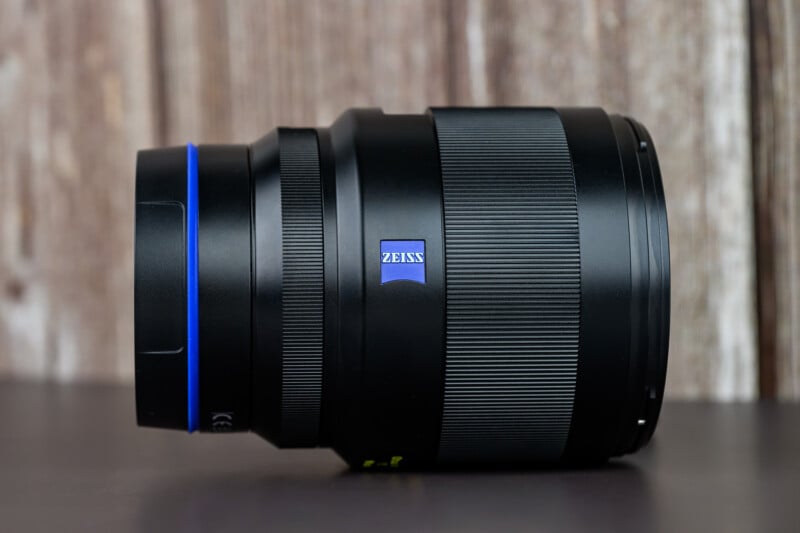 A black Zeiss camera lens with a blue accent ring and the Zeiss logo sits on a dark surface against a blurred wooden background.