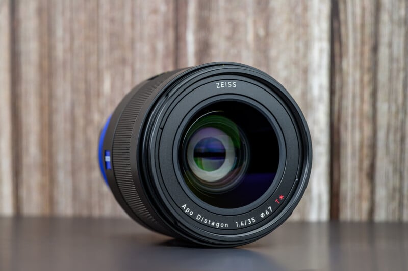 A close-up of a Zeiss camera lens, model Apo Distagon 1.4/35, placed vertically on a dark surface with a wooden background. The lens glass reflects light, and branding details are clearly visible.