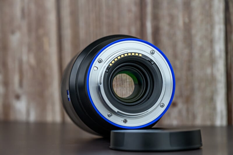 A close-up of a camera lens facing backward, showing the electronic contacts and rear glass element. The lens sits on a dark surface with its rear lens cap removed and placed beside it. The background is blurred wood paneling.