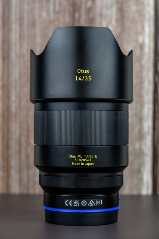 A Zeiss Otus 1.4/35 camera lens with a lens hood, standing upright on a wooden surface with a blurred wooden background. Yellow and white text with specifications is visible on the lens barrel.