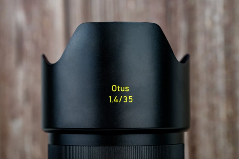 A close-up of a black camera lens hood featuring yellow text that reads "Otus 1.4/35" against a blurred wooden background.