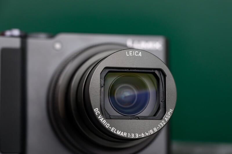 A close-up of a Leica camera lens with "LEICA" and technical details inscribed on the rim, attached to a black camera body against a blurred green background.