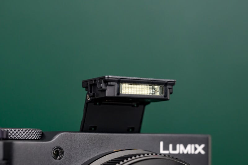 Close-up of a black Lumix camera with the built-in flash unit popped up against a plain green background.