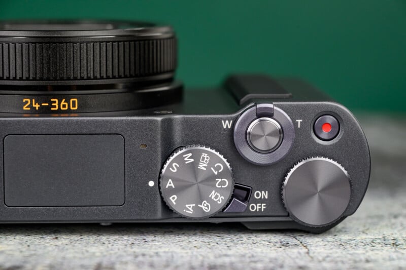 Close-up of a digital camera’s top, showing a zoom lever, mode dial with various settings, power switch, and part of the lens extending from the left. The background is green and the surface is textured gray.