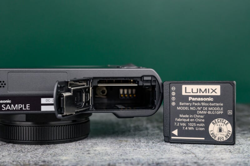 A close-up of a camera with its battery compartment open, next to a Panasonic Lumix DMW-BLG10PP rechargeable lithium-ion battery standing upright on a gray surface.