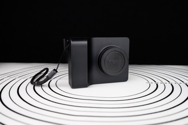 A compact black digital camera with a lens cap labeled "Micro" is placed on a white surface with black concentric circles, against a dark background. The camera has a wrist strap attached on the left side.