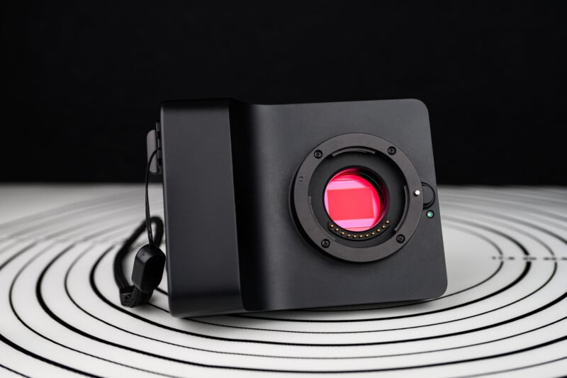A black digital camera body with a visible red image sensor is placed on a white surface with black concentric circles, against a solid black background.