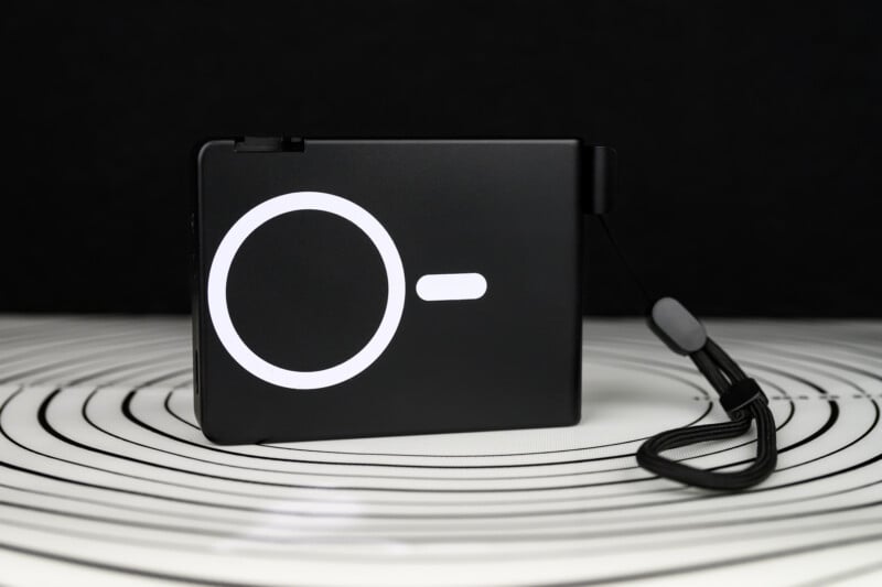 A black rectangular electronic device with a white circular and line design on the front, standing upright on a white surface with concentric black circles, and a black wrist strap attached to the side.
