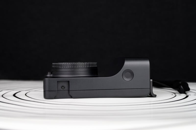 A black digital camera without a lens, viewed from the side, is placed on a white surface with black concentric circles. The background is solid black.