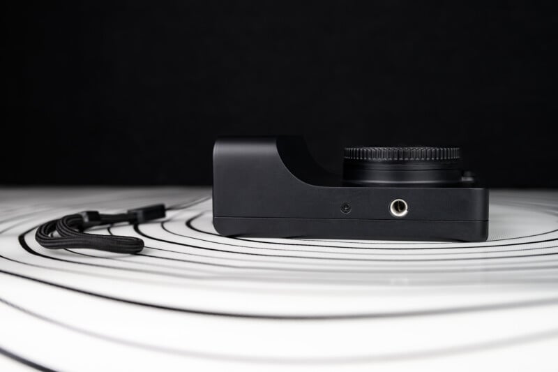 A black digital camera body with no lens attached sits on a white, circular patterned surface, next to a detached black camera strap, against a dark background.