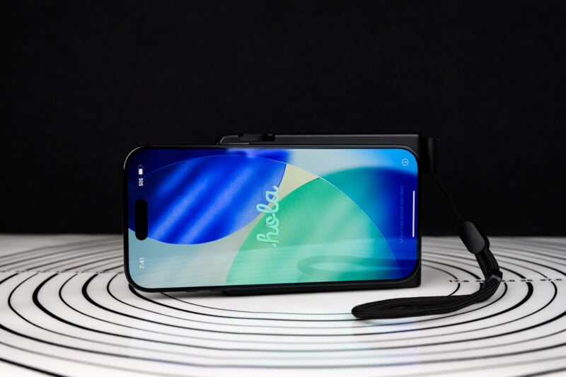 A smartphone with a colorful "hola" welcome screen is lying horizontally on a white surface with black circular lines, against a black background. A black strap is attached to the phone case.