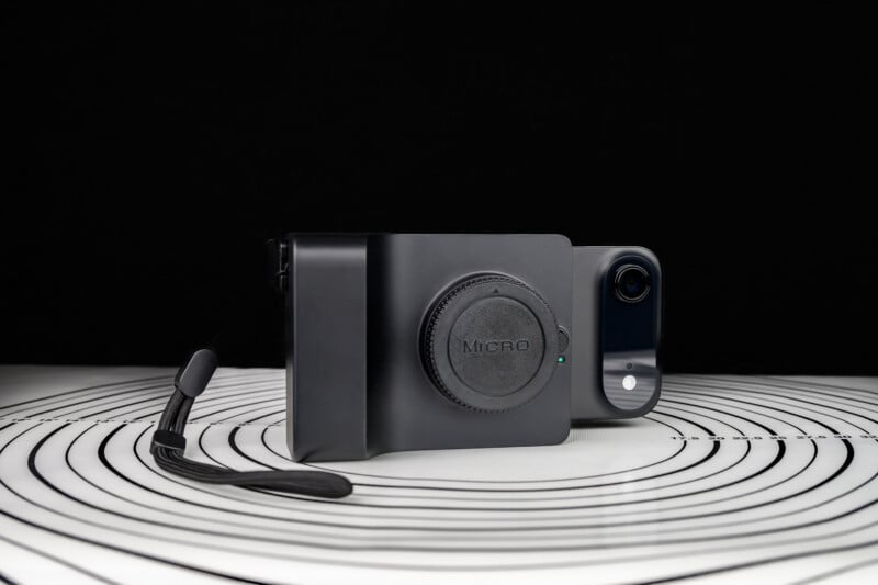 A smartphone in a black camera grip case with a lens cap labeled "Micro," placed on a white surface with concentric black circles, and a black strap attached, against a black background.