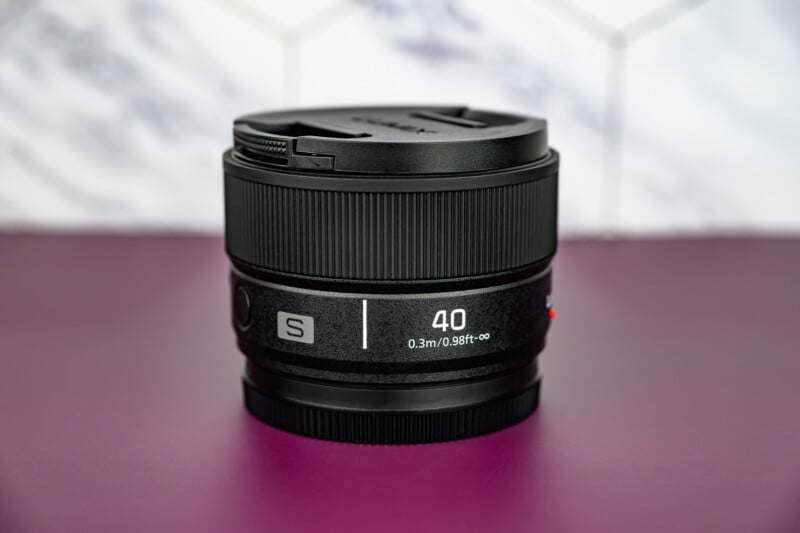 A black camera lens with "S 40 0.3m/0.98ft-∞" labeled on it sits on a purple surface, with a blurred white and gray patterned background.