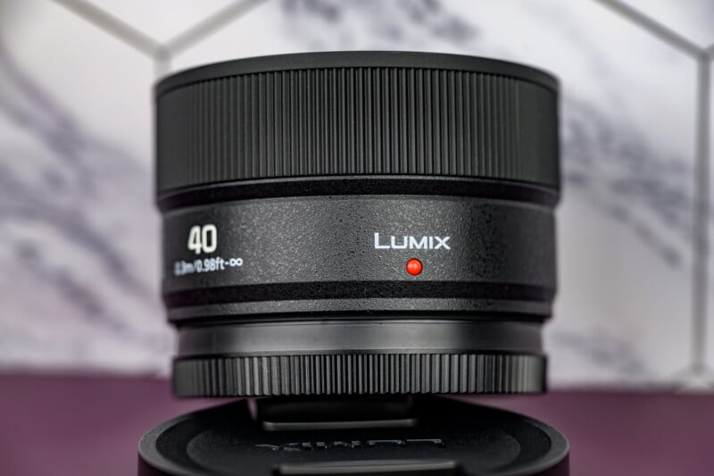 Close-up of a black Lumix camera lens with "40" and "LUMIX" written on it, placed on a lens cap against a blurred white and purple background with geometric patterns.