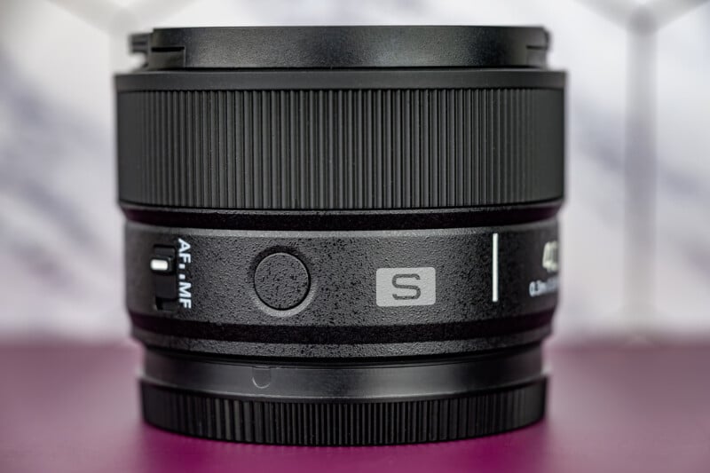 Close-up of a black camera lens with focus and mode switches, and "S" logo visible. The background is blurred with purple and white tones. The lens is resting on a purple surface.