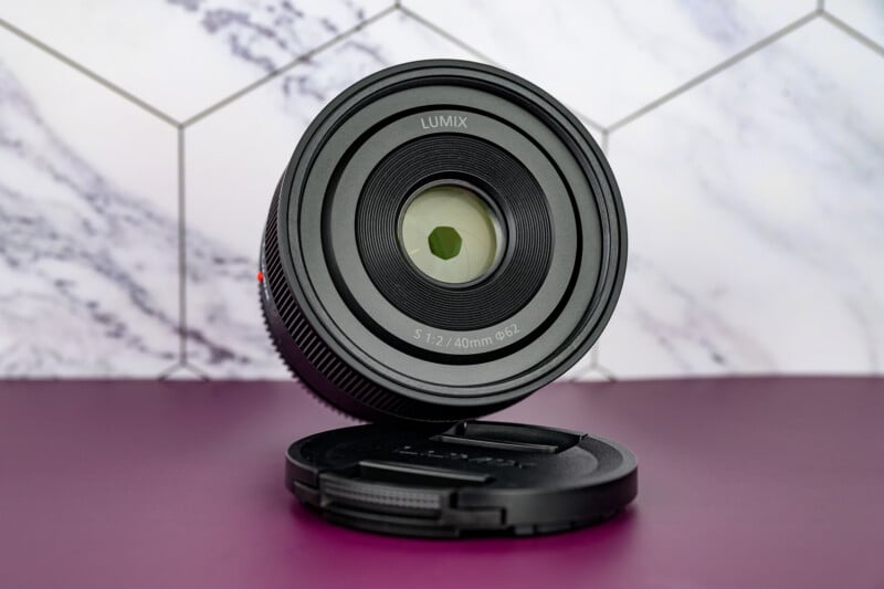 A Lumix camera lens stands upright with its lens cap lying flat in front of it, set on a purple surface against a white and gray geometric background.