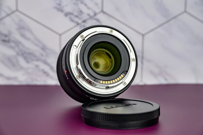 A camera lens stands upright with its rear glass and electronic contacts visible, next to a detached Lumix lens cap, on a purple surface with a hexagonal tile background.